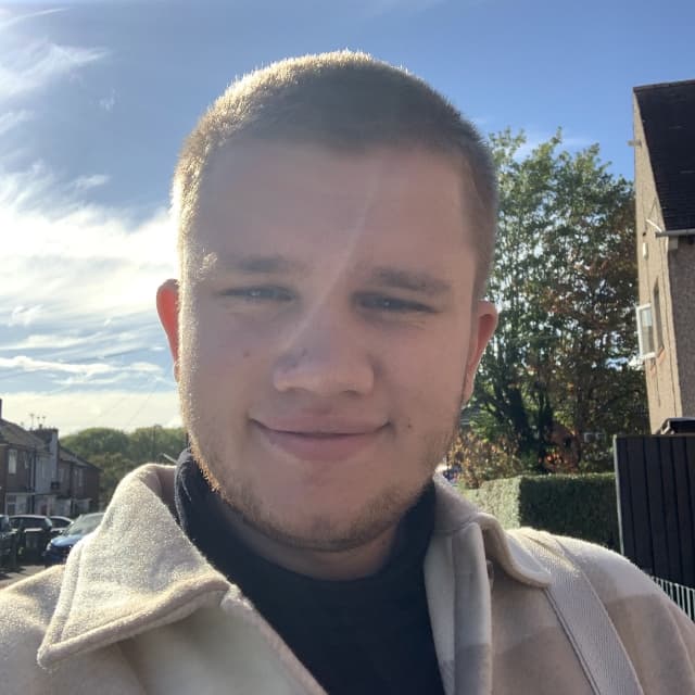 My personal photo headshot(selfie) - I'm smiling confidently at the camera, while wearing beige coat. You can also spot some buildings and trees in the background.